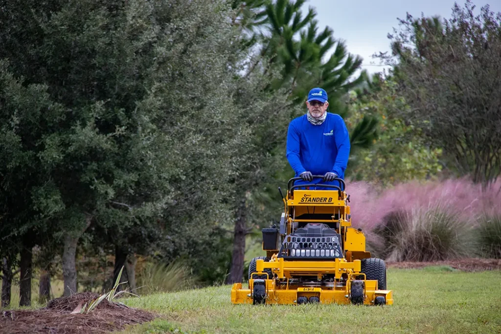 Grounds crew member in a blue shirt and cap rides a yellow stand-on mower, cutting grass in a landscaped area with trees and shrubs.