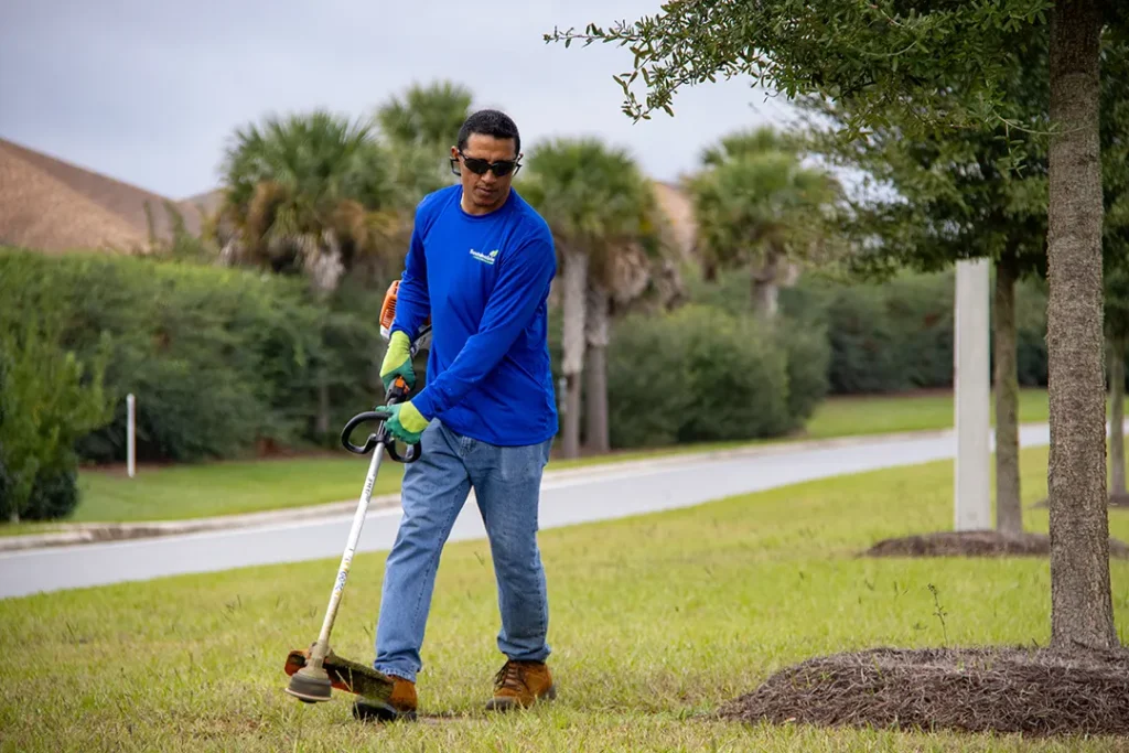 A landscaper wearing sunglasses uses a weed trimmer on a grassy verge beside a neighborhood street with palm trees in the background.