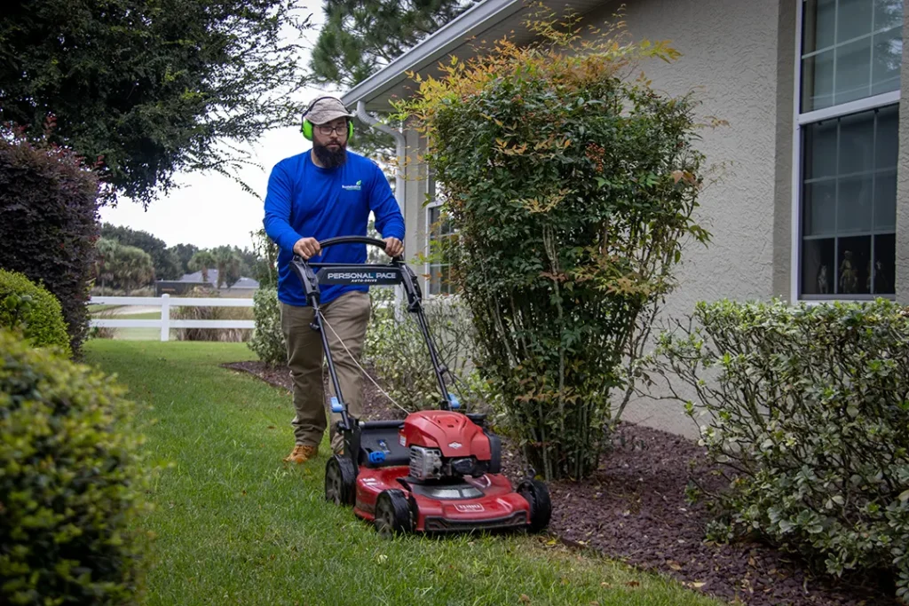 Worker in a blue shirt and ear protection pushes a red lawn mower along a narrow strip of grass beside a house and shrubs.