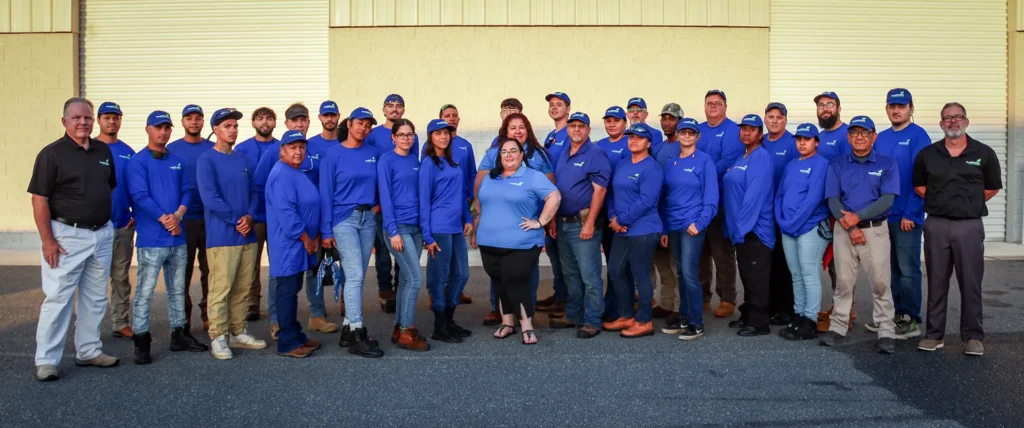 Company team photo: A diverse crew wearing blue long-sleeve shirts and branded hats gathers for a group portrait outside.