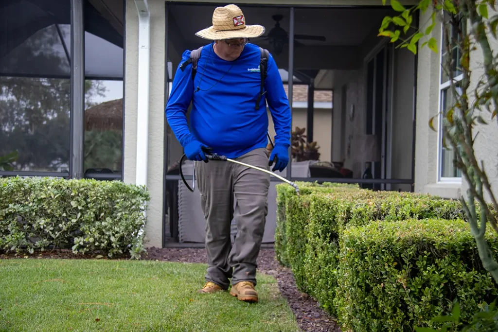 Worker in a blue long-sleeve shirt and straw hat uses a spray wand along neatly trimmed hedges beside a home.