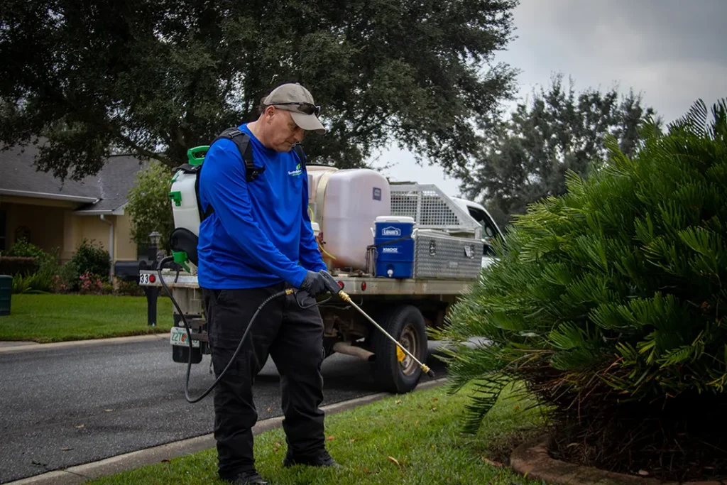 Grounds maintenance worker in a blue shirt and cap sprays a large shrub with a wand sprayer, with a service truck and tank parked behind.