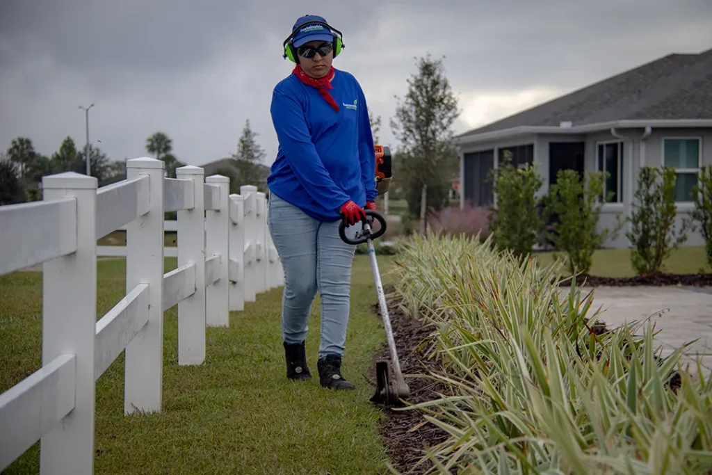 Worker in a blue uniform, ear protection, and red gloves uses a string trimmer to edge a mulched planting bed beside a white fence.