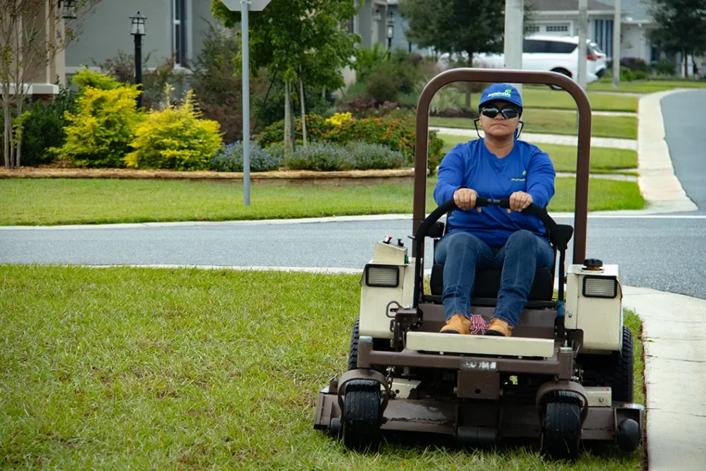 Grounds crew member in a blue shirt and cap rides a lawn mower, cutting grass in a residential area with trees and shrubs.