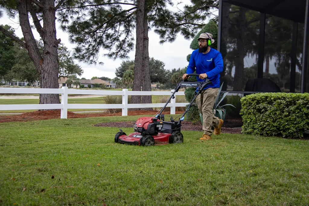 Worker in a blue shirt and ear protection pushes a red lawn mower along a narrow strip of grass beside a white fence.